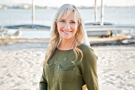 Dr. Melanie Parker standing on a sandy beach with water and sailboats visible in the background.