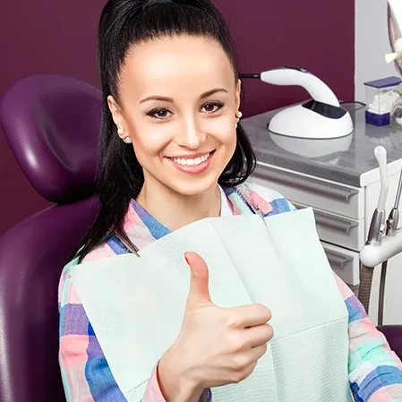 Person seated in a dental chair holding a dental bib and giving a thumbs-up, with equipment nearby.
