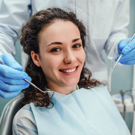 Person seated in a dental chair while gloved hands hold dental instruments nearby.